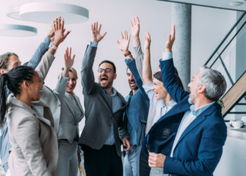 Shot of group of business people celebrating their success in the office. Multiracial group of corporate business persons on a business meeting in board room. Cheerful business team raising their hands together in the air.
