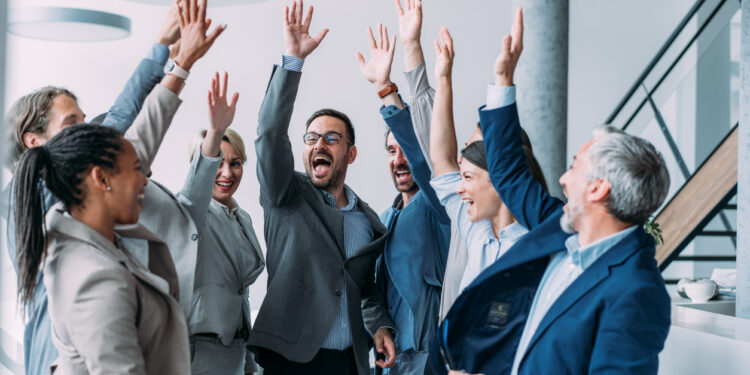 Shot of group of business people celebrating their success in the office. Multiracial group of corporate business persons on a business meeting in board room. Cheerful business team raising their hands together in the air.