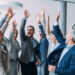 Shot of group of business people celebrating their success in the office. Multiracial group of corporate business persons on a business meeting in board room. Cheerful business team raising their hands together in the air.